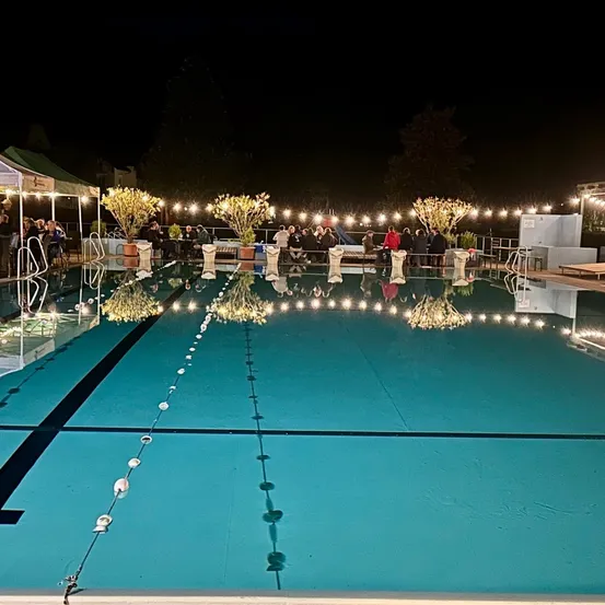 Eine nächtliche Szene eines Pools mit Menschen, die sich umher versammelt haben, beleuchtet von Lichterketten und Topfpflanzen.