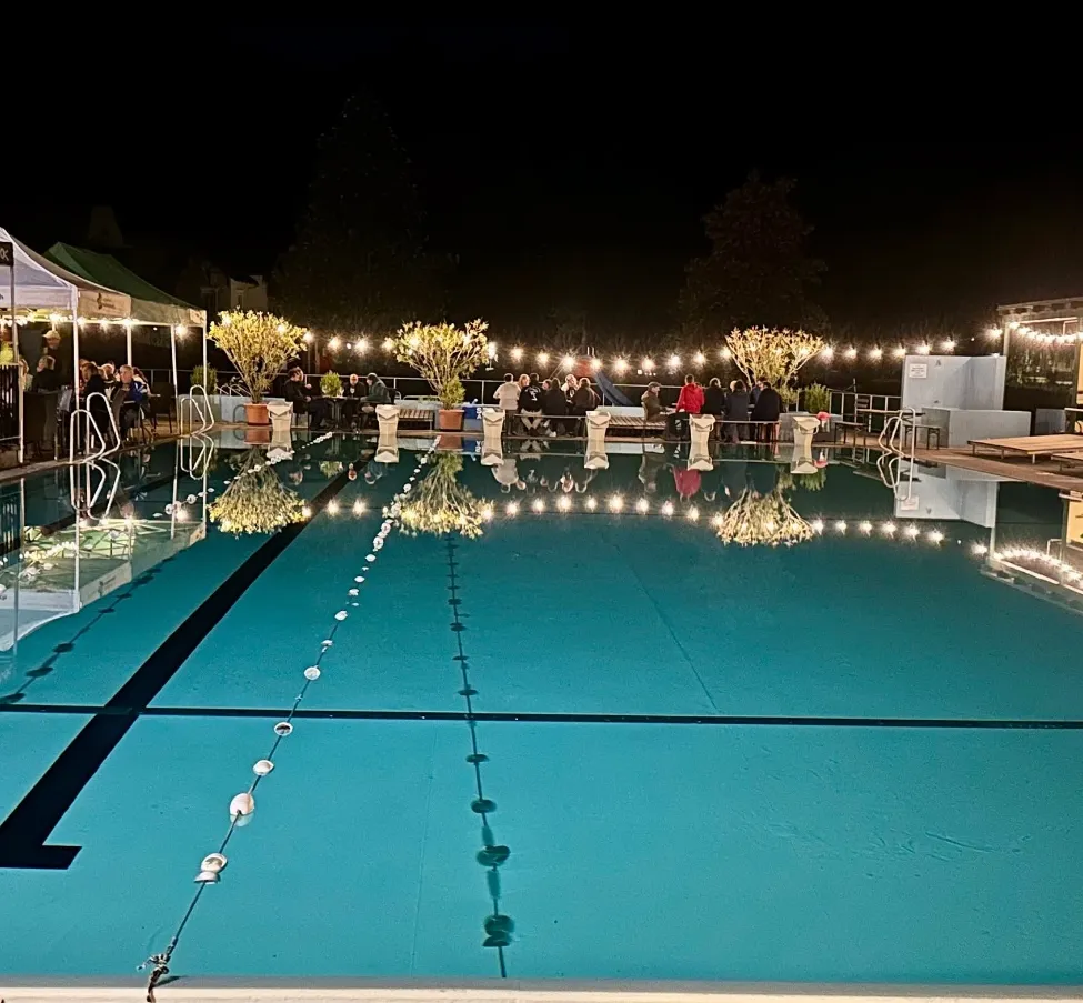 Eine nächtliche Szene eines Pools mit Menschen, die sich umher versammelt haben, beleuchtet von Lichterketten und Topfpflanzen.