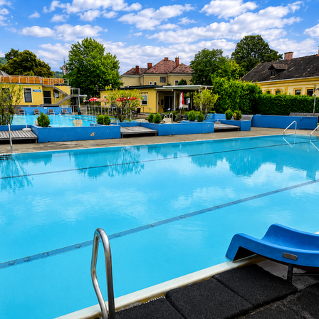 Ein großer Außenpool mit blauem Wasser, umgeben von gelben Gebäuden und viel Grün. Eine blaue Rutsche befindet sich auf der rechten Seite des Pools.