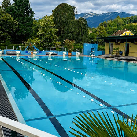 Ein Freibad mit blauem Wasser, umgeben von einer Terrasse, Liegestühlen und einem Gebäude mit gelbem Dach. Im Hintergrund befinden sich Bäume und Berge.