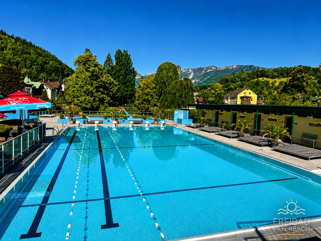 Ein Freibad mit blauem Wasser und klarem Himmel. Im Hintergrund befinden sich Bäume und Berge.