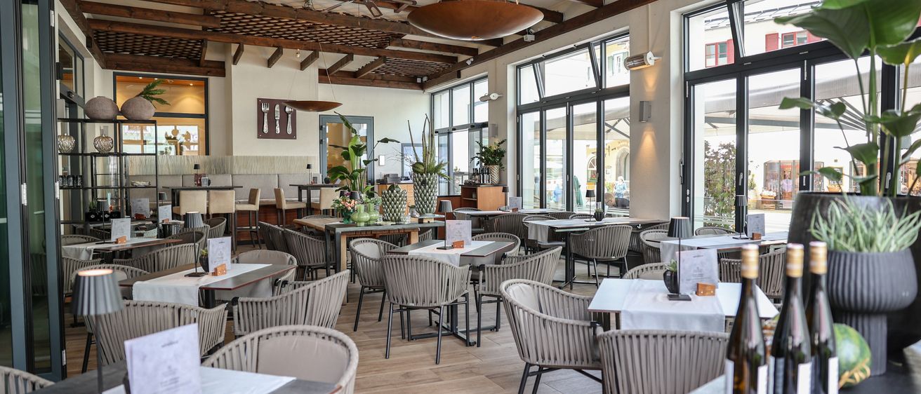 Ein Innenrestaurant mit Holzböden, hohen Decken und großen Fenstern. Tische und Stühle sind aufgestellt, mit Pflanzen und Menükarten auf den Tischen. Es hängen Ventilatoren und Lampen von der Decke.