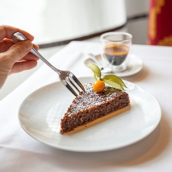 Eine Hand hält eine Gabel, bereit, ein Stück Schokoladenkuchen mit einer kleinen Orange obenauf zu essen, begleitet von einem kleinen Glas Kaffee auf einem weißen Tisch.