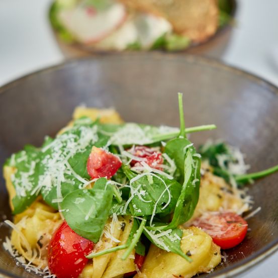 Eine Nahaufnahme eines Tellers mit Nudelsalat mit Tomaten, Spinat und geriebenem Käse. Ein weiteres Gericht ist im Hintergrund zu sehen.