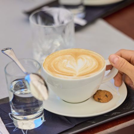 Eine Hand hält eine Tasse mit Herz-Design-Latte, begleitet von einem Glas Wasser und einem Keks auf einer Untertasse.