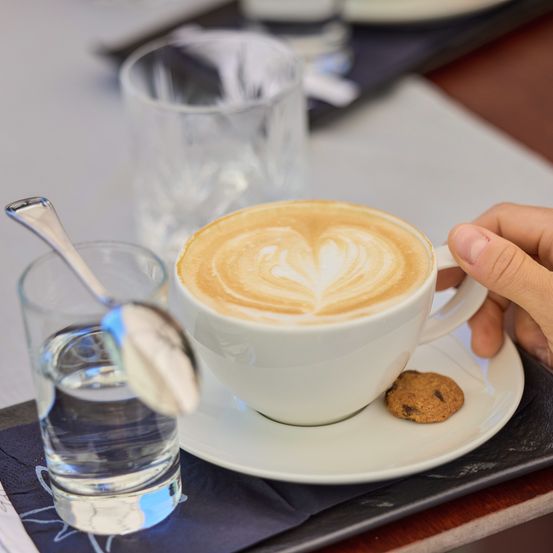 Eine Hand hält eine Tasse mit Herz-Design-Latte, begleitet von einem Glas Wasser und einem Keks auf einer Untertasse.