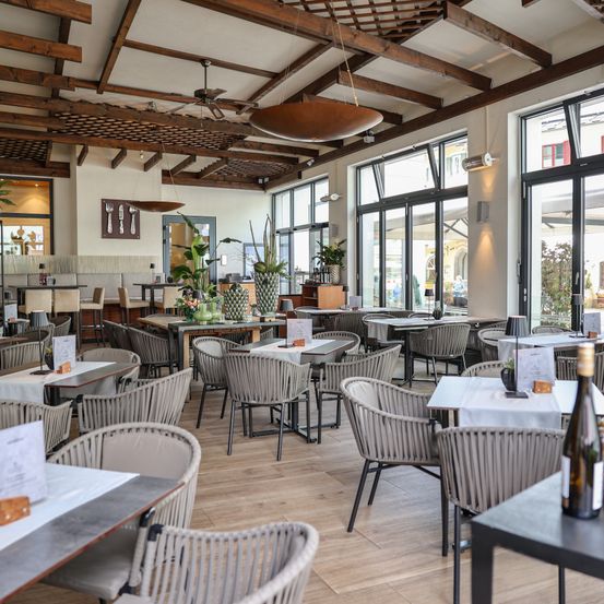 Ein Restaurant-Innenraum mit Holzboden, großen Fenstern und Deckenventilatoren. Tische und Stühle sind in Reihen angeordnet, einige Tische haben Vasen und Menüs. Ein Barbereich ist links sichtbar.