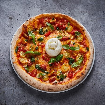 Eine ganze Pizza mit Kirschtomaten, Basilikumblättern und einem Mozzarella-Ball, serviert auf einem weißen Teller vor grauem Hintergrund.