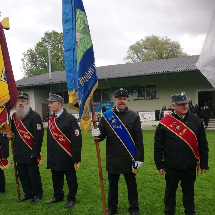 Vier Männer in schwarzen Uniformen mit roten Schärpen stehen auf einem Feld und halten Fahnen, mit einem Gebäude und Menschen im Hintergrund.