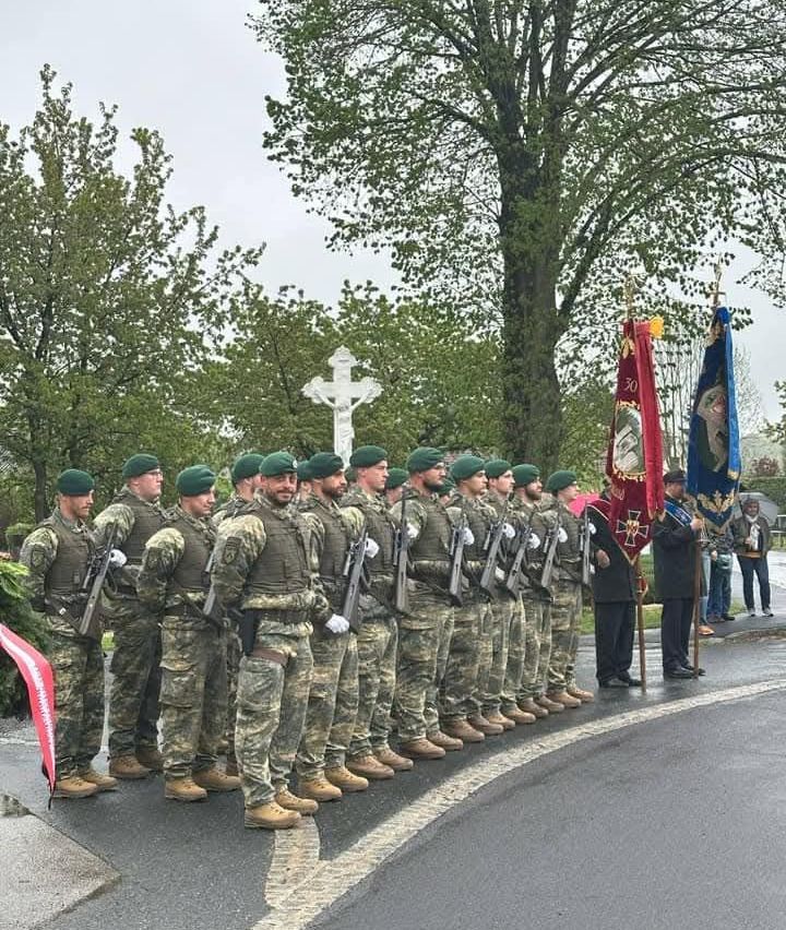 Eine Gruppe von Soldaten in Tarnuniformen und grünen Mützen steht in einer Reihe, hält Gewehre und steht vor einem Kreuz.