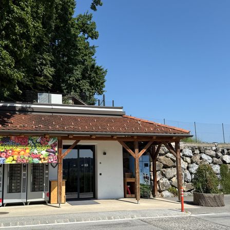 Ein Laden mit braunem Dach und einem Schild, das lokale Produkte anbietet. Der Laden hat Glastüren und eine kleine Veranda. Zwei Schließfächer zum Aufbewahren von Gegenständen befinden sich auf der linken Seite.
