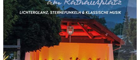 Ein nächtliches Konzert mit klassischer Musik, Lichtshow und Feuerwerk am Rathausplatz. Das Salonorchester Bad Goisern spielt auf einer von warmen Lichtern erleuchteten Bühne. Zuschauer sitzen auf Bänken unter den Bäumen.