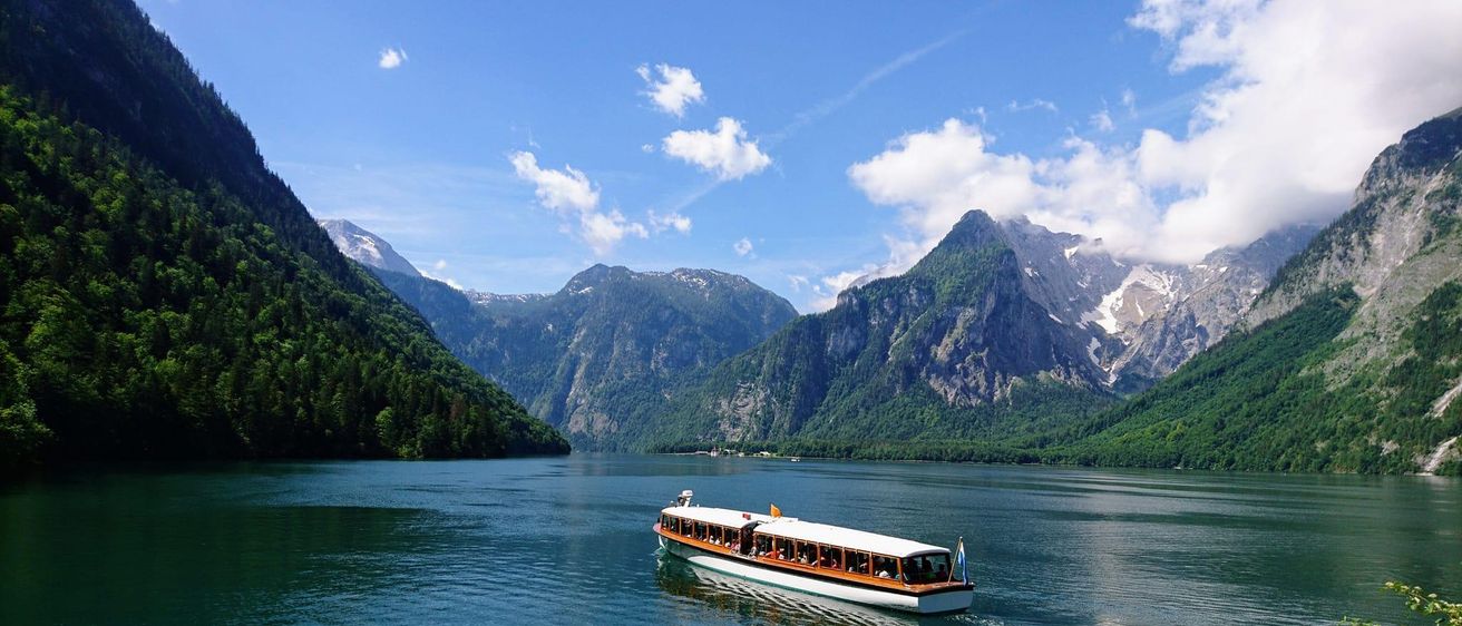 Ein Boot fährt auf einem ruhigen See mit Bergen und Grünflächen im Hintergrund. Das Boot ist weiß mit braunen Verzierungen und hat mehrere Personen an Bord.