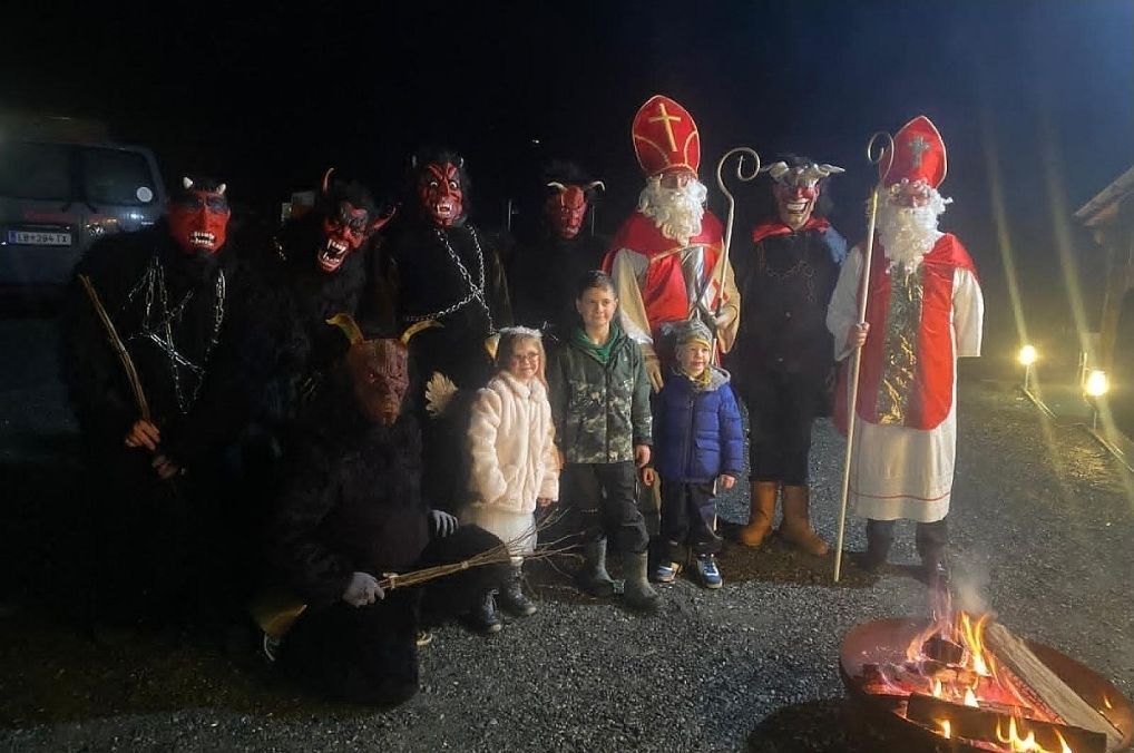 Eine Gruppe von Menschen in Kostümen posiert um ein Lagerfeuer. Einige tragen Teufelsmasken, während andere als Weihnachtsmann gekleidet sind. Vor ihnen stehen einige Kinder.