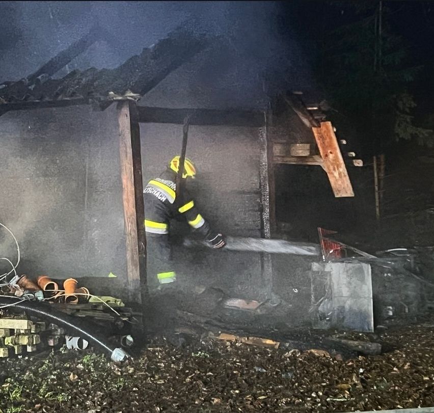 Ein Feuerwehrmann sprüht Wasser auf ein ausgebranntes Gebäude mit beschädigtem Dach und Wänden. Trümmerteile sind umher verstreut, und Rauch erfüllt die Luft.