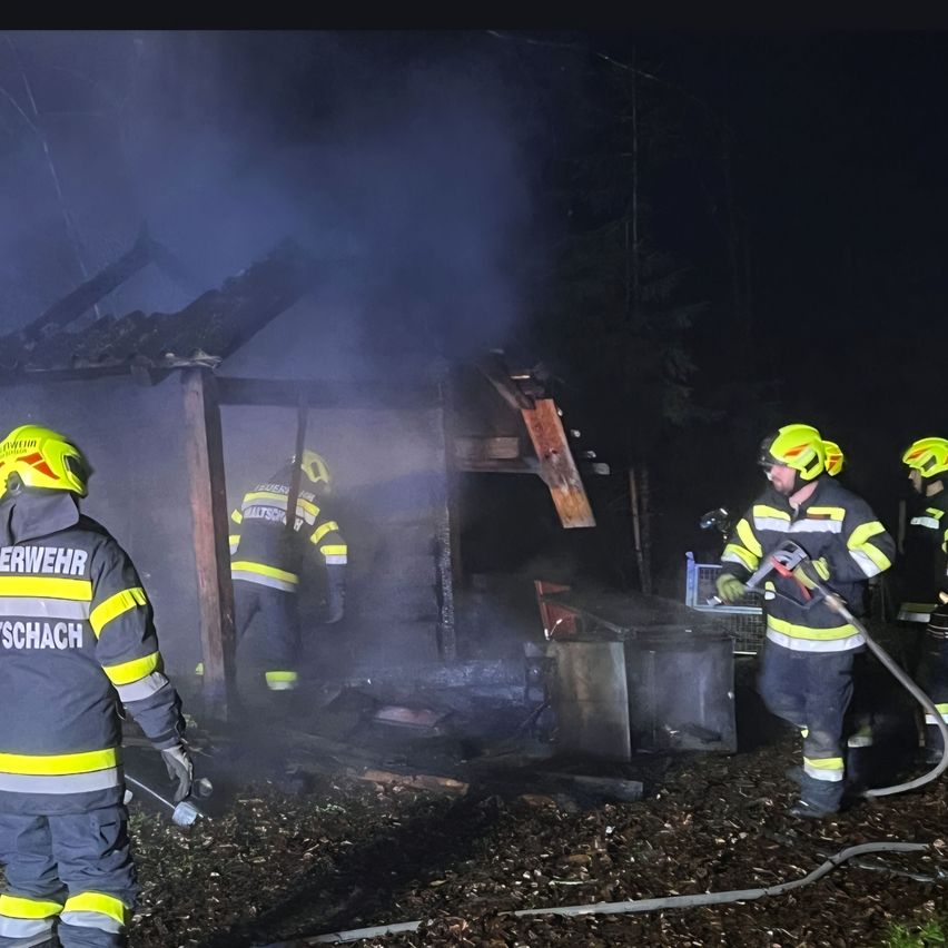 Feuerwehrleute in gelben und schwarzen Uniformen bekämpfen einen Brand in einer Holzstruktur und verwenden einen Schlauch, um das Feuer zu löschen. Die Szene ist bei Nacht mit Bäumen im Hintergrund.