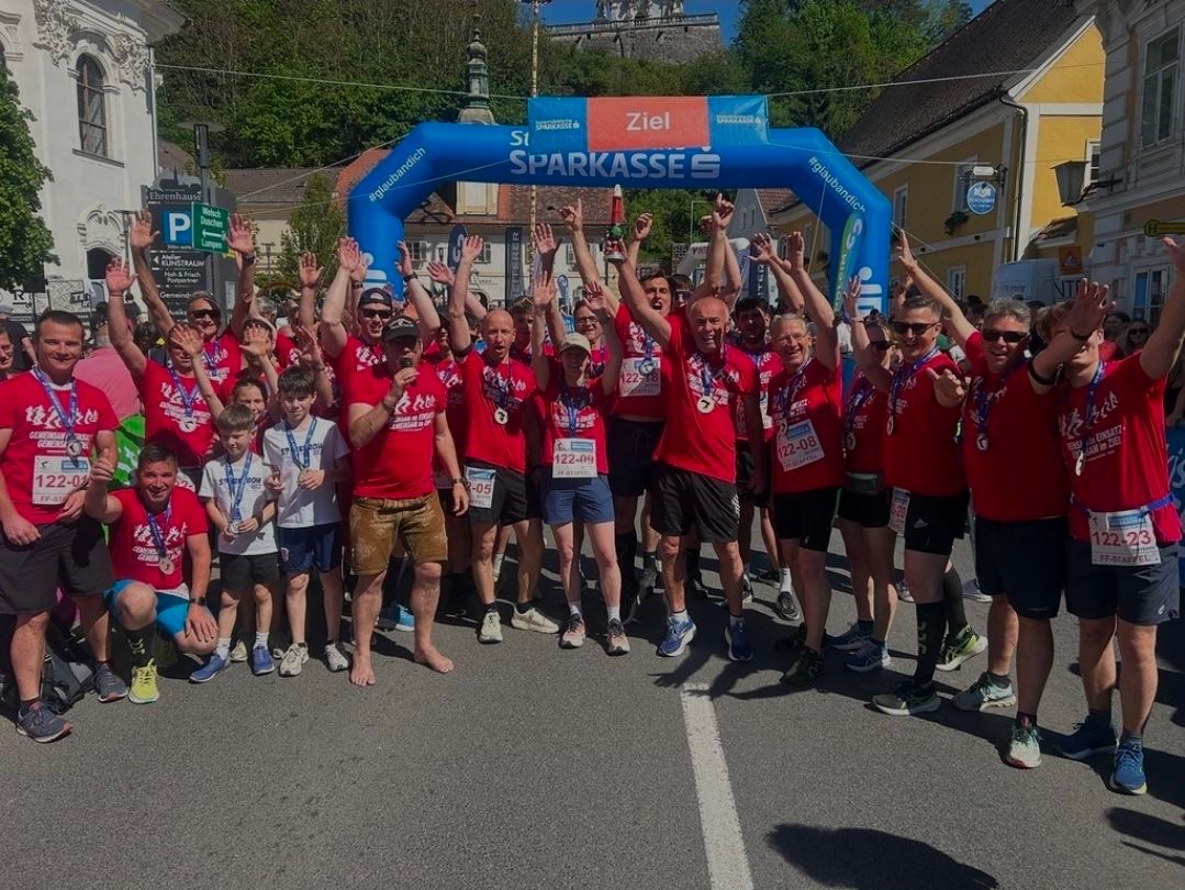 Runners in red shirts celebrate at the finish line of a marathon, holding medals and cheering, under a blue arch.