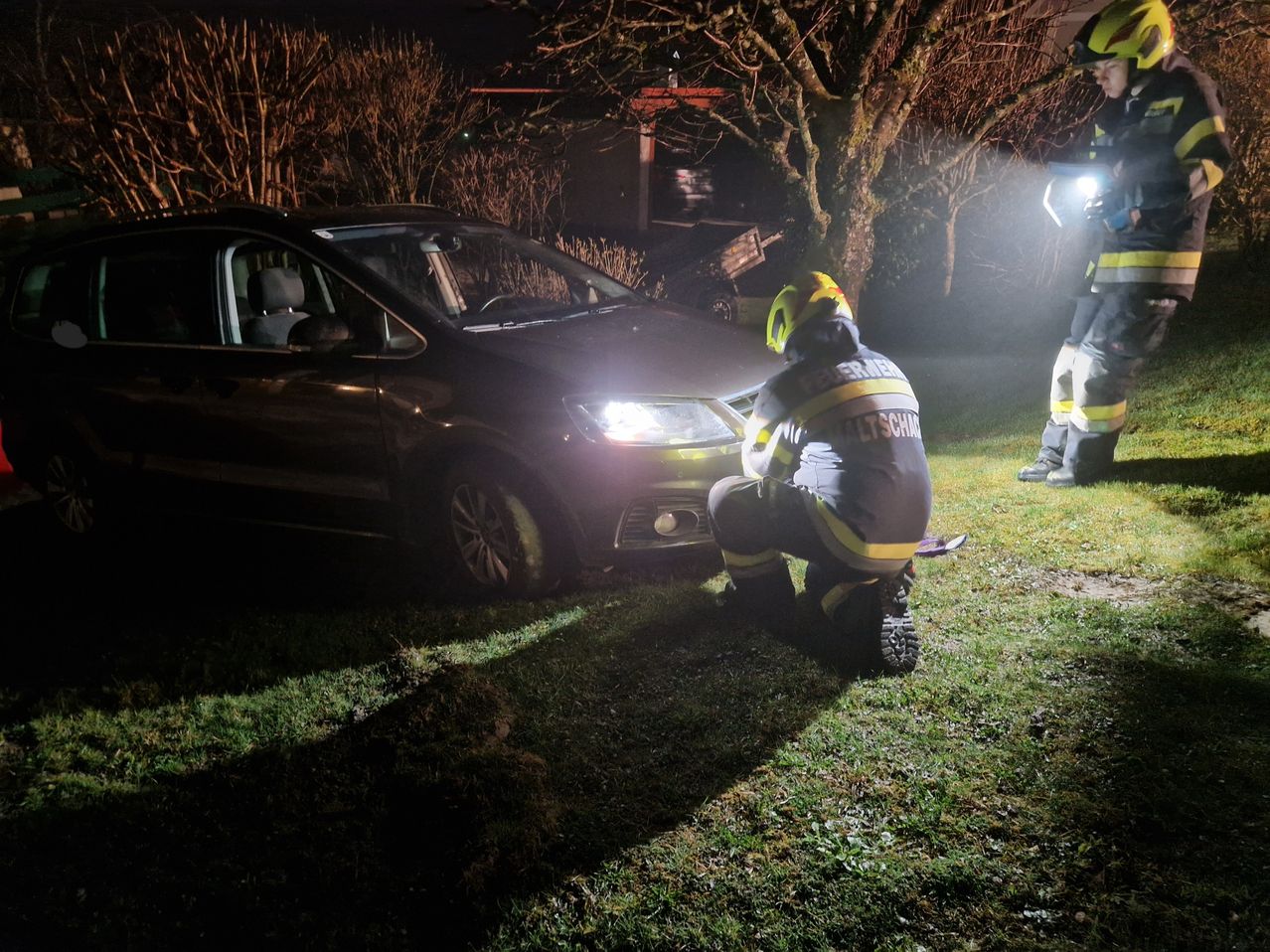 Zwei Feuerwehrleute inspizieren ein beschädigtes Fahrzeug bei Nacht unter einem hellen Licht.