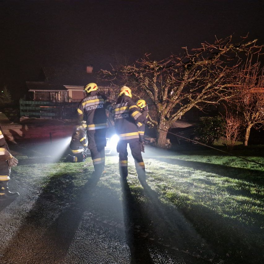 Feuerwehrleute arbeiten bei Nacht an einem Autounfall. Einer kniet und die anderen stehen mit Taschenlampen. Bäume und ein Haus sind im Hintergrund.