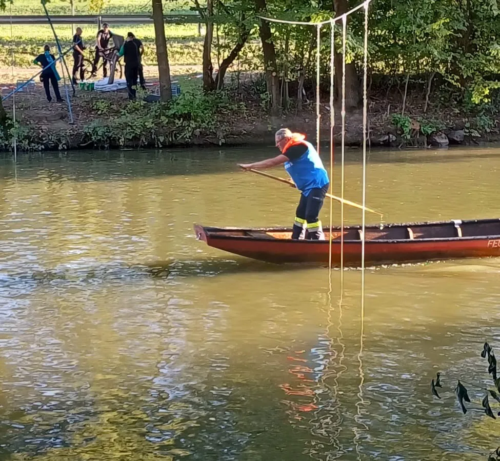 Ein Mann in einem blauen Shirt und einer Rettungsweste rudert mit einem Boot auf einem Fluss, während Leute am Ufer stehen.
