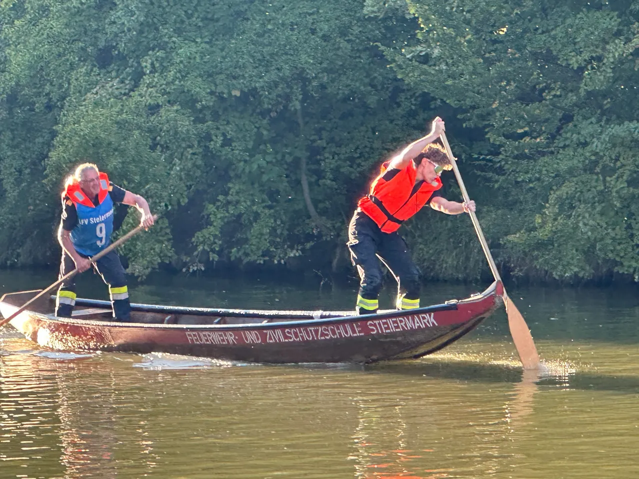 Zwei Männer in Rettungswesten paddeln ein rotes Boot mit der Aufschrift 'Feuerwehr und Zivilschutzschule'.