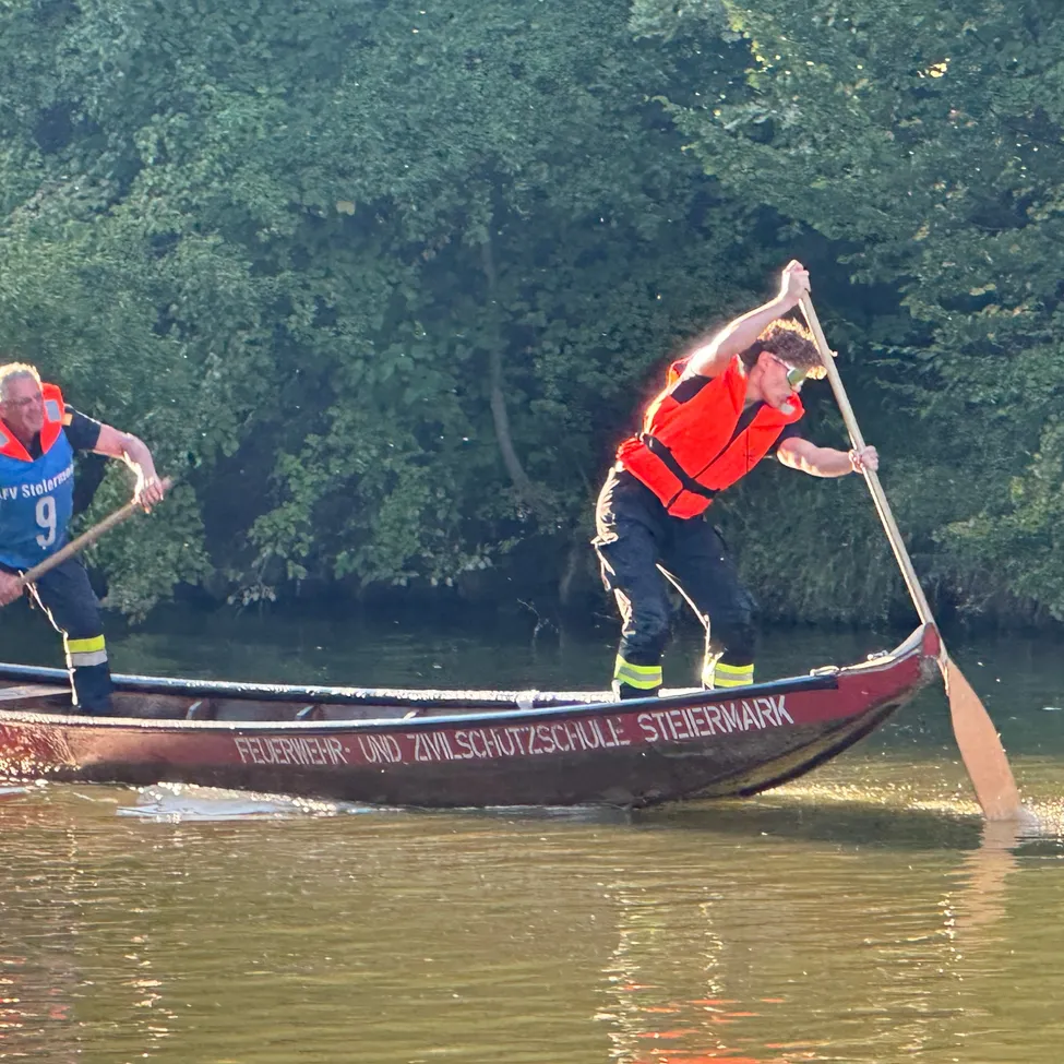 Zwei Männer in Rettungswesten paddeln ein rotes Boot mit der Aufschrift 'Feuerwehr und Zivilschutzschule'.