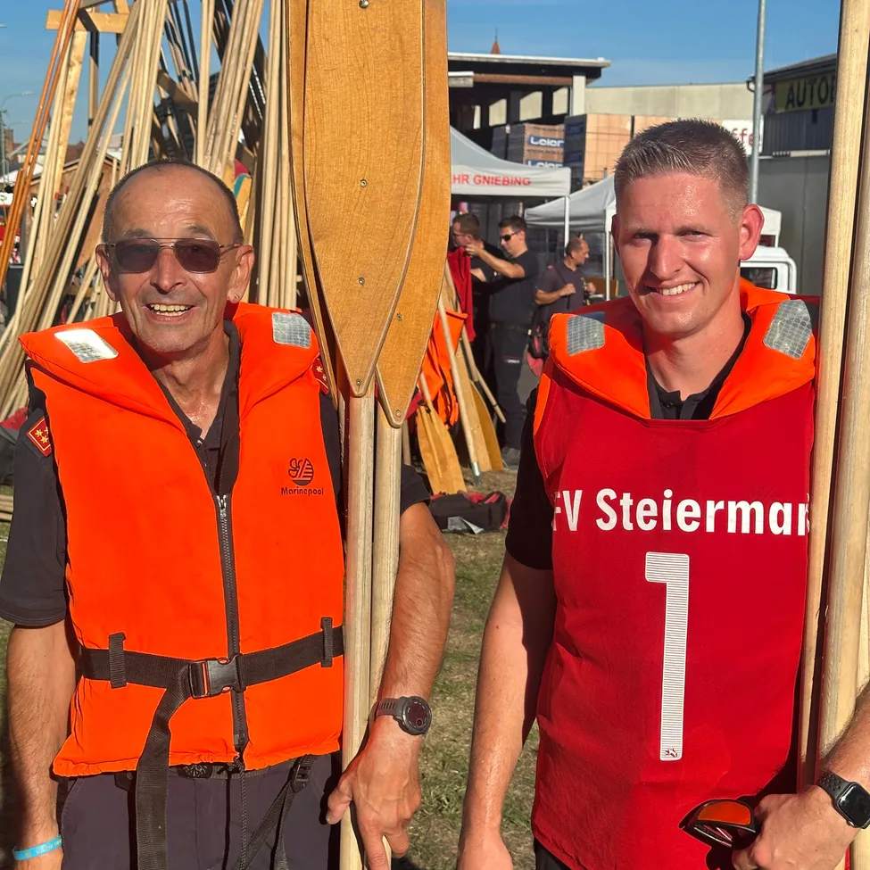 Zwei Männer in orangenen Rettungswesten, einer hält ein Paddel, lächeln für ein Foto. Dahinter ein Stapel von Paddeln und ein verschwommener Hintergrund mit Menschen.