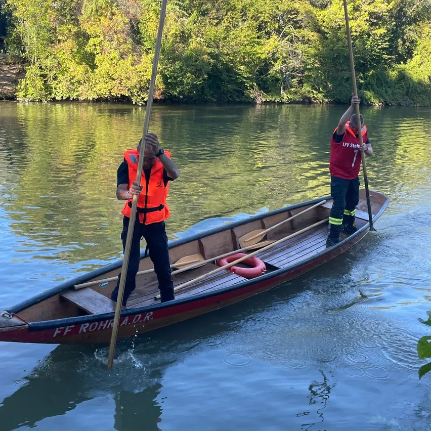 Zwei Personen in Rettungswesten paddeln ein Boot auf einem Fluss mit Bäumen im Hintergrund.