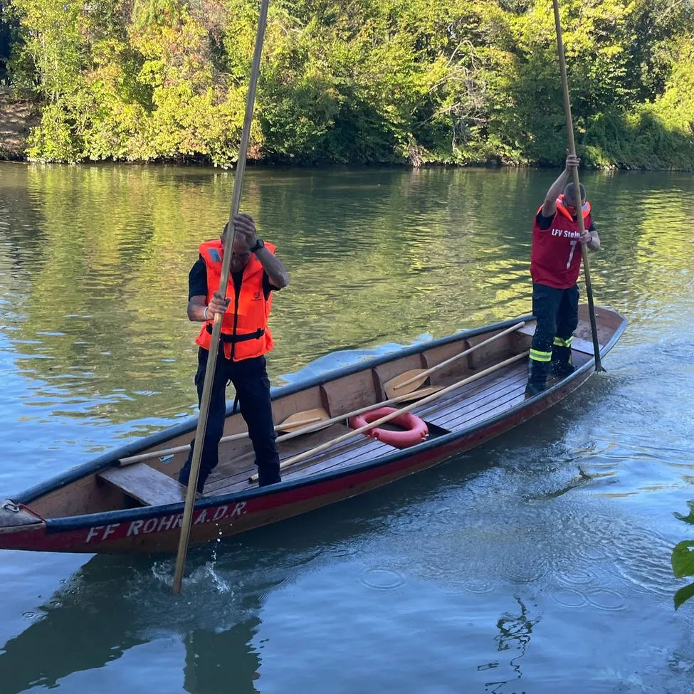 Zwei Personen in Rettungswesten paddeln ein Boot auf einem Fluss mit Bäumen im Hintergrund.