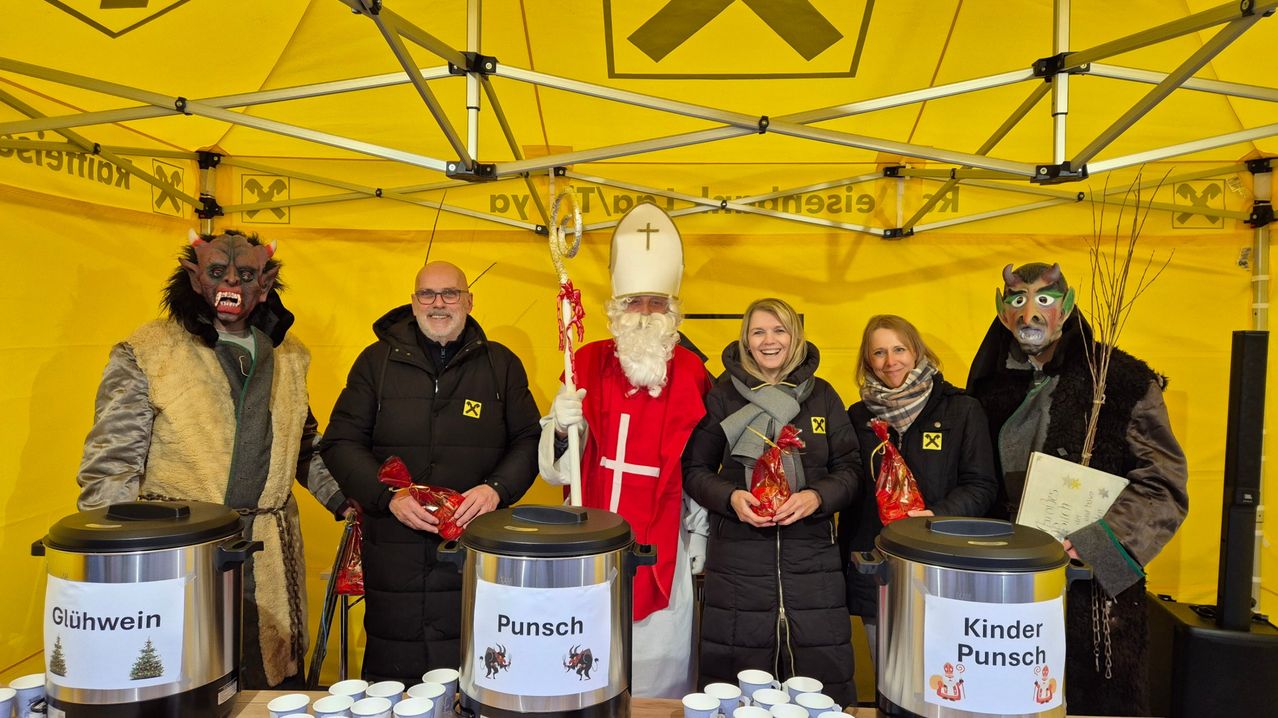 Eine Gruppe von Menschen unter einem gelben Zelt. Eine Person ist als Heiliger gekleidet. Die anderen halten Geschenke und lächeln. Getränkebehälter stehen auf dem Tisch.