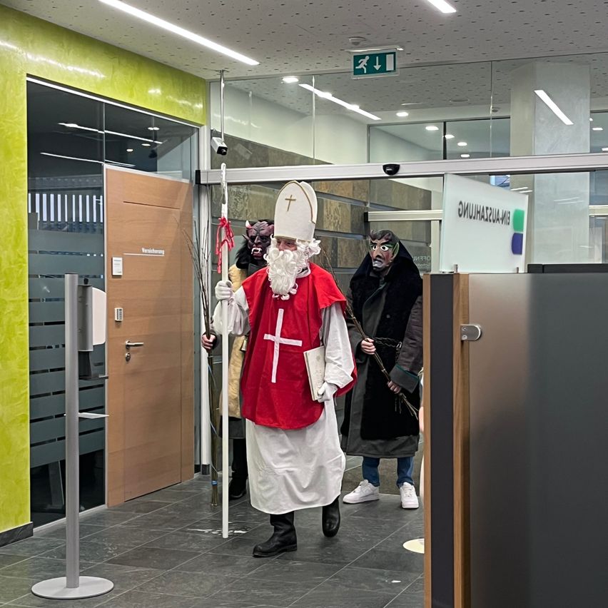 Eine als Bischof verkleidete Person geht durch einen Korridor und hält einen Stab. Dahinter folgen zwei Personen in Masken. Der Korridor hat Glaswände, eine Holztür und ein Schild.