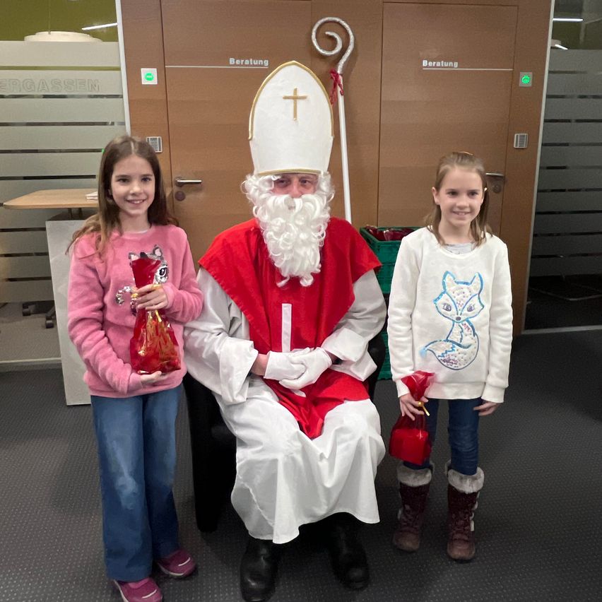 Zwei junge Mädchen stehen neben einem Mann, der als Weihnachtsmann verkleidet ist, in einem Raum mit einer braunen Tür, die mit 'Beratuung' beschriftet ist. Die Mädchen halten kleine rote Tüten.