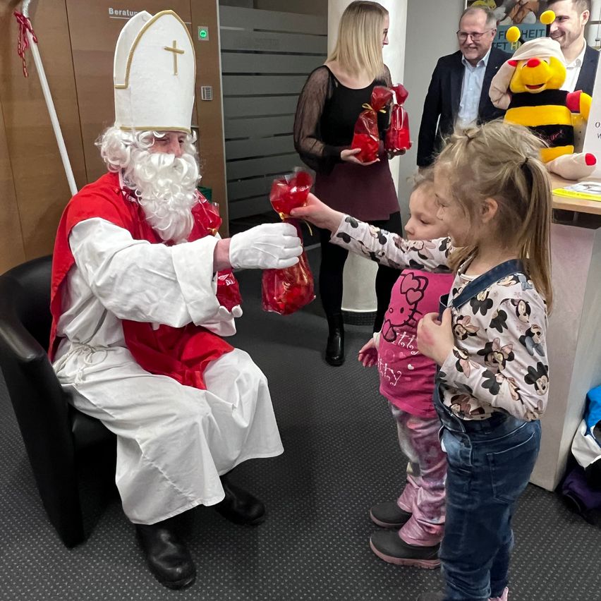 Ein Mann, der als Weihnachtsmann verkleidet ist, sitzt auf einem Stuhl und gibt einem kleinen Mädchen ein Geschenk, während eine Frau und ein Biesteddy dahinter stehen.