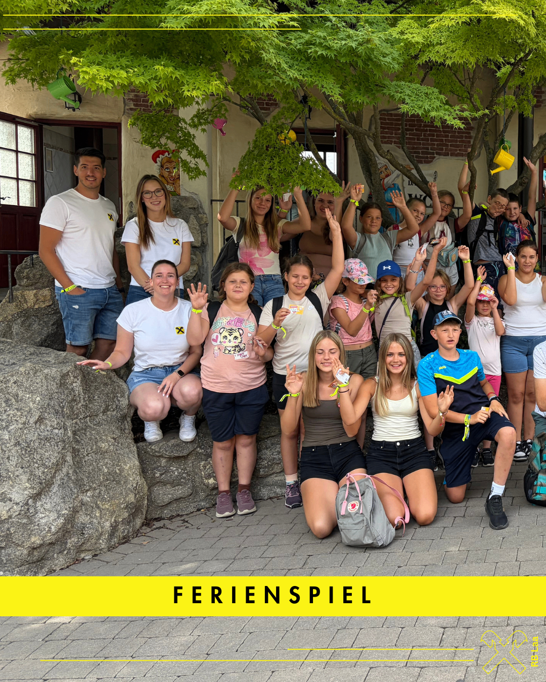 Eine Gruppe von Menschen, einschließlich Erwachsener und Kindern, posiert für ein Foto vor einem Gebäude. Sie alle lächeln und winken mit den Händen. Die Erwachsenen tragen weiße T-Shirts, und die Kinder tragen verschiedene Outfits. Das Bild hat ein Wasserzeichen, das Ferienspiel lautet.