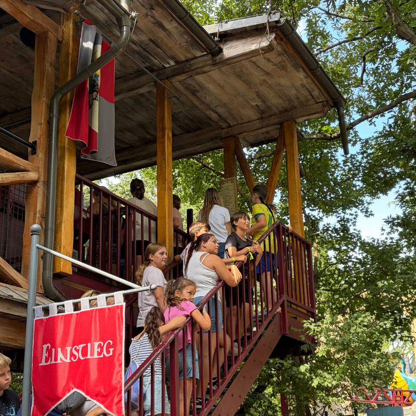 Eine Gruppe von Menschen versammelt sich auf einer Holztreppe, möglicherweise in einem Park. Einige schauen nach unten, andere nach oben. Eine rote Fahne mit weißem Text befindet sich auf der linken Seite.