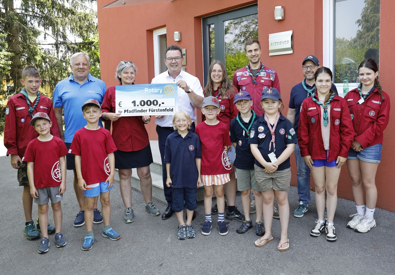 Eine Gruppe von Erwachsenen und Kindern posiert für ein Foto vor einem Gebäude. Die Erwachsenen halten einen Scheck über 1.000 Euro für Pfadfinder Furstenfeld.