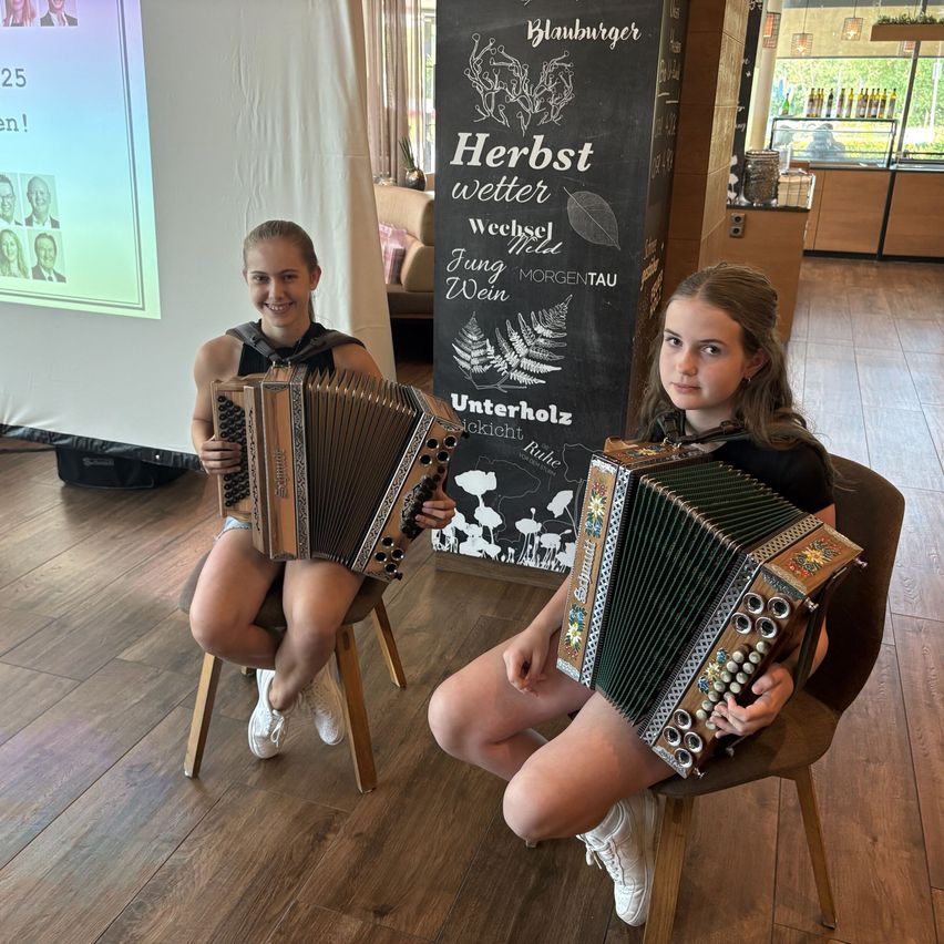 Zwei junge Mädchen sitzen auf Stühlen und spielen Akkordeons. Hinter ihnen steht eine Tafel mit dem Text Herbstwetter. Der Boden ist aus Holz, und links befindet sich ein weißer Vorhang.