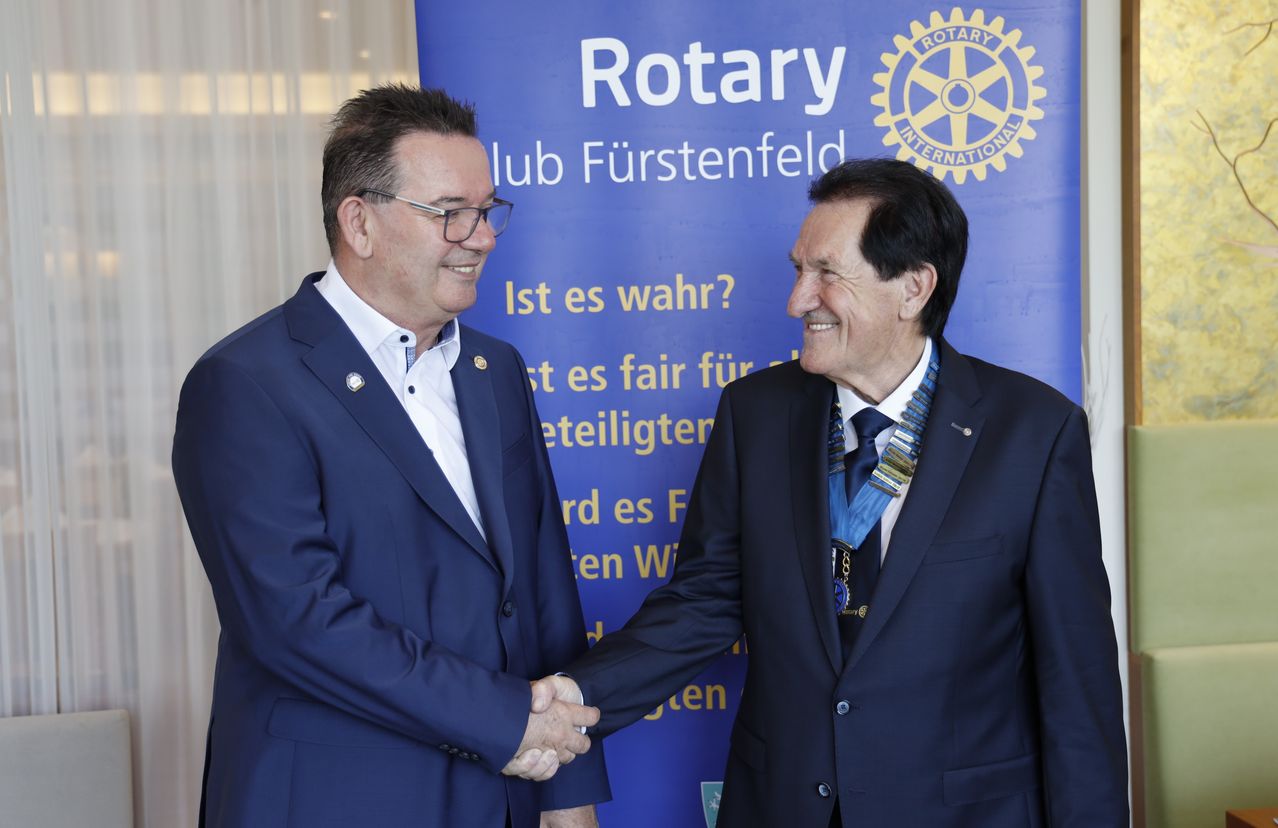 Zwei Männer in Anzügen lächeln und schütteln sich die Hände vor einem Rotary Club-Banner.