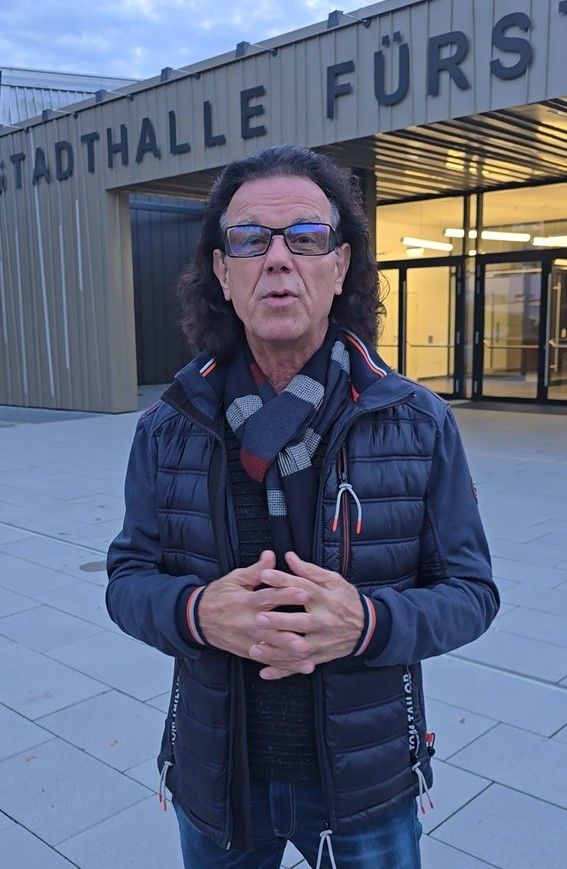Ein Mann mit langen Haaren und einer Brille steht vor einem Gebäude namens Stadthalle Furo. Er trägt eine blaue Jacke und einen Schal. Er hält die Hände zusammengeklappt.