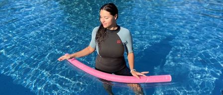 Eine Frau steht in einem Schwimmbecken, hält eine rosa Schwimmhilfe und hat die Augen geschlossen.