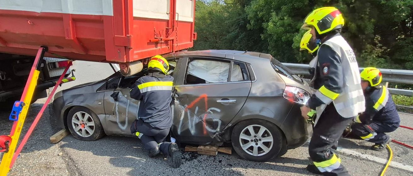 Zwei Feuerwehrleute in gelben Helmen arbeiten an einem demoliertn Auto unter einem roten LKW am Straßenrand mit Graffiti darauf.