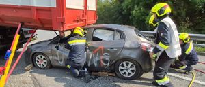 Zwei Feuerwehrleute in gelben Helmen arbeiten an einem demoliertn Auto unter einem roten LKW am Straßenrand mit Graffiti darauf.