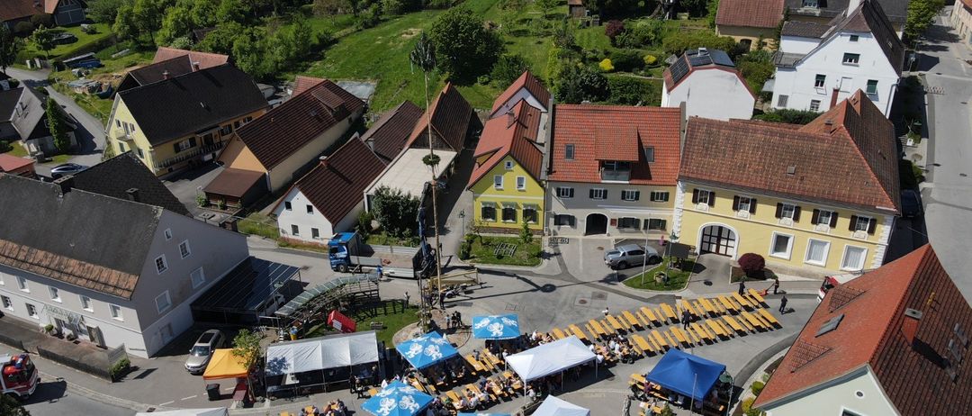 Luftaufnahme eines Dorfes mit mehreren Häusern und Gebäuden. Ein Straßenmarkt mit Zelten und Menschen ist in der Mitte. Bäume und Grünflächen umgeben das Gebiet, mit Bergen in der Ferne.