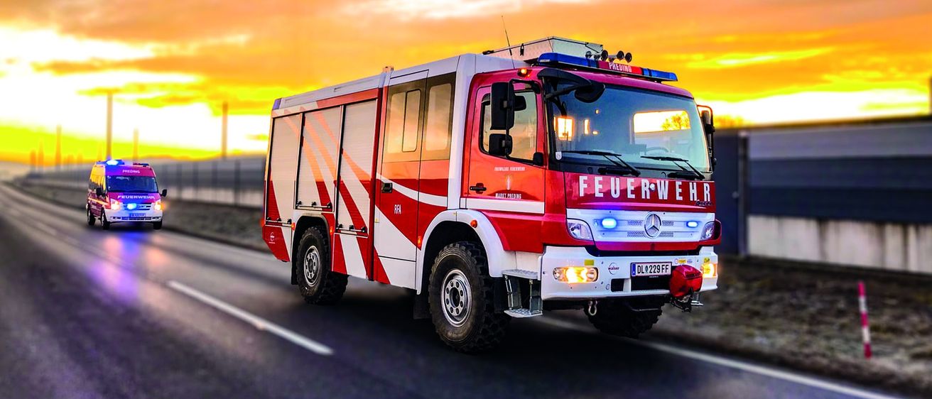 Ein roter Feuerwehrwagen mit blauen Lichtern fährt auf einer Straße mit Sonnenuntergang im Hintergrund. Ein weiteres Fahrzeug ist dahinter.