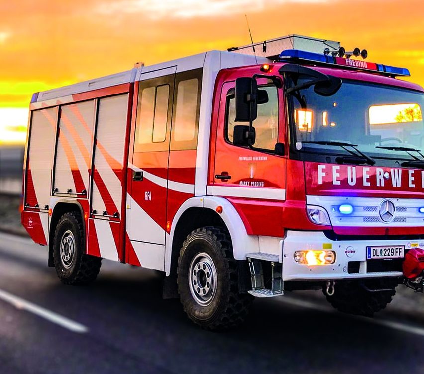 Ein roter Feuerwehrwagen mit blauen Lichtern fährt auf einer Straße mit Sonnenuntergang im Hintergrund. Ein weiteres Fahrzeug ist dahinter.
