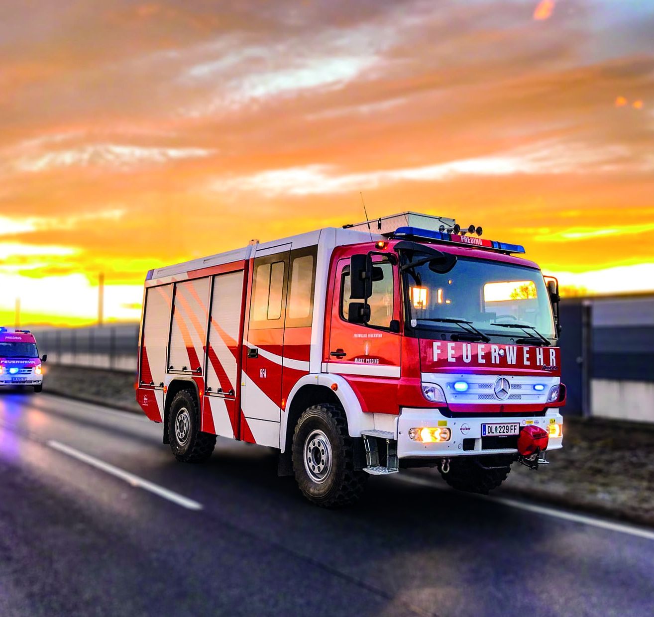 Ein roter und weißer Feuerwehrwagen fährt auf der Straße, während die Sonne im Hintergrund untergeht. Das Fahrzeug hat das Wort 'Feuerwehr' auf der Front geschrieben.