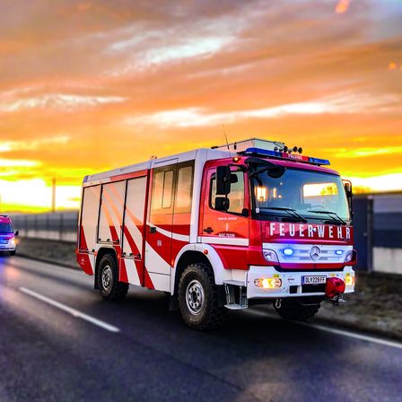 Ein roter und weißer Feuerwehrwagen fährt auf der Straße, während die Sonne im Hintergrund untergeht. Das Fahrzeug hat das Wort 'Feuerwehr' auf der Front geschrieben.