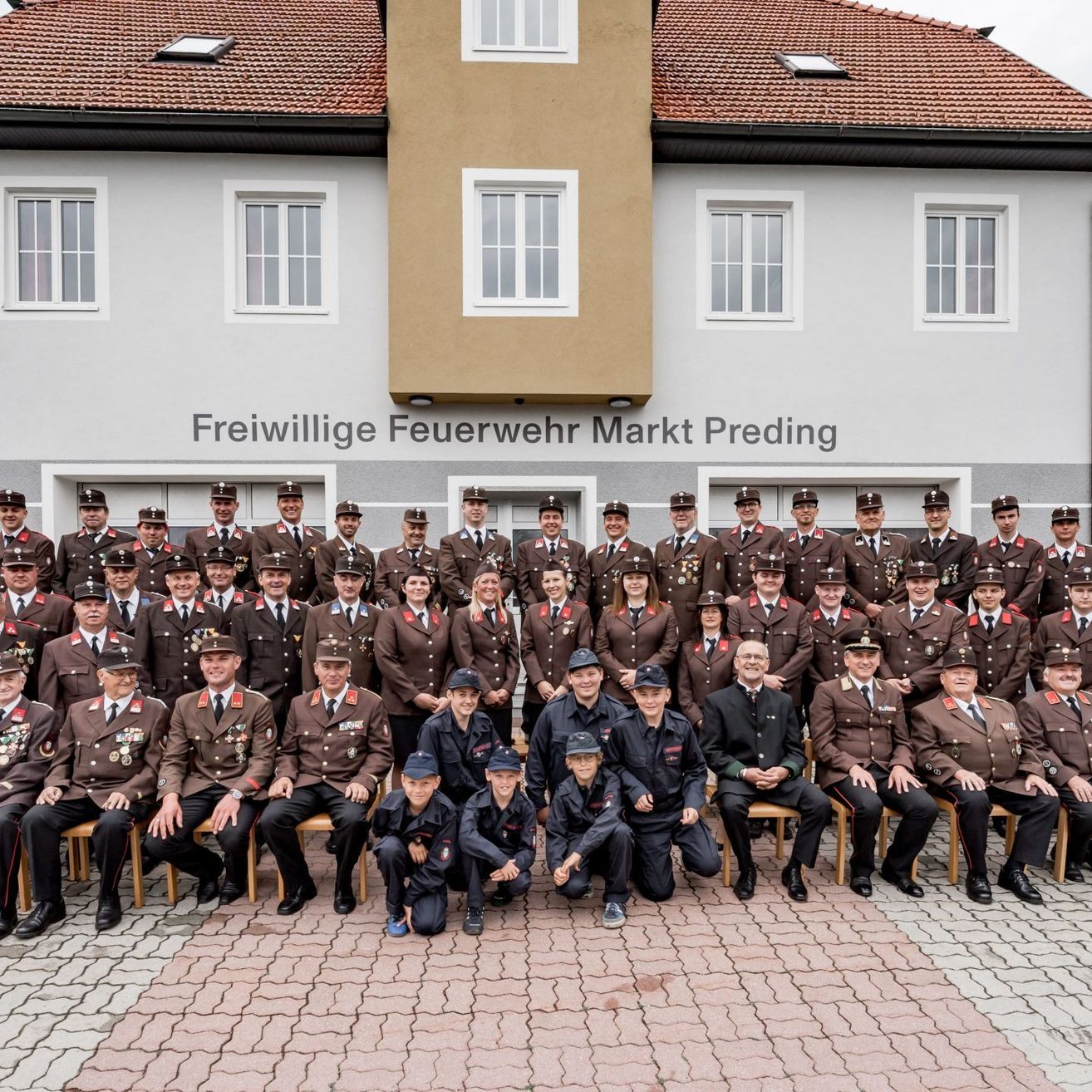 Eine große Gruppe von Freiwilligen der Freiwillige Feuerwehr Markt Preding posiert für ein Foto vor ihrem Feuerwehrhaus. Sie sind alle in Uniform gekleidet, einige sitzen auf Stühlen, andere stehen. Das Gebäude hat mehrere Fenster und ein rotes Dach.