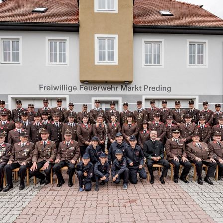 Eine große Gruppe von Freiwilligen der Freiwillige Feuerwehr Markt Preding posiert für ein Foto vor ihrem Feuerwehrhaus. Sie sind alle in Uniform gekleidet, einige sitzen auf Stühlen, andere stehen. Das Gebäude hat mehrere Fenster und ein rotes Dach.