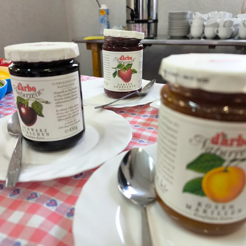 Zwei Gläser mit Marmelade stehen auf einem Tisch mit rot-weißem Tischtuch. Ein Glas hat ein Erdbeeretikett und das andere ein Orangenetikett. Löffel und Untertassen sind ebenfalls vorhanden.